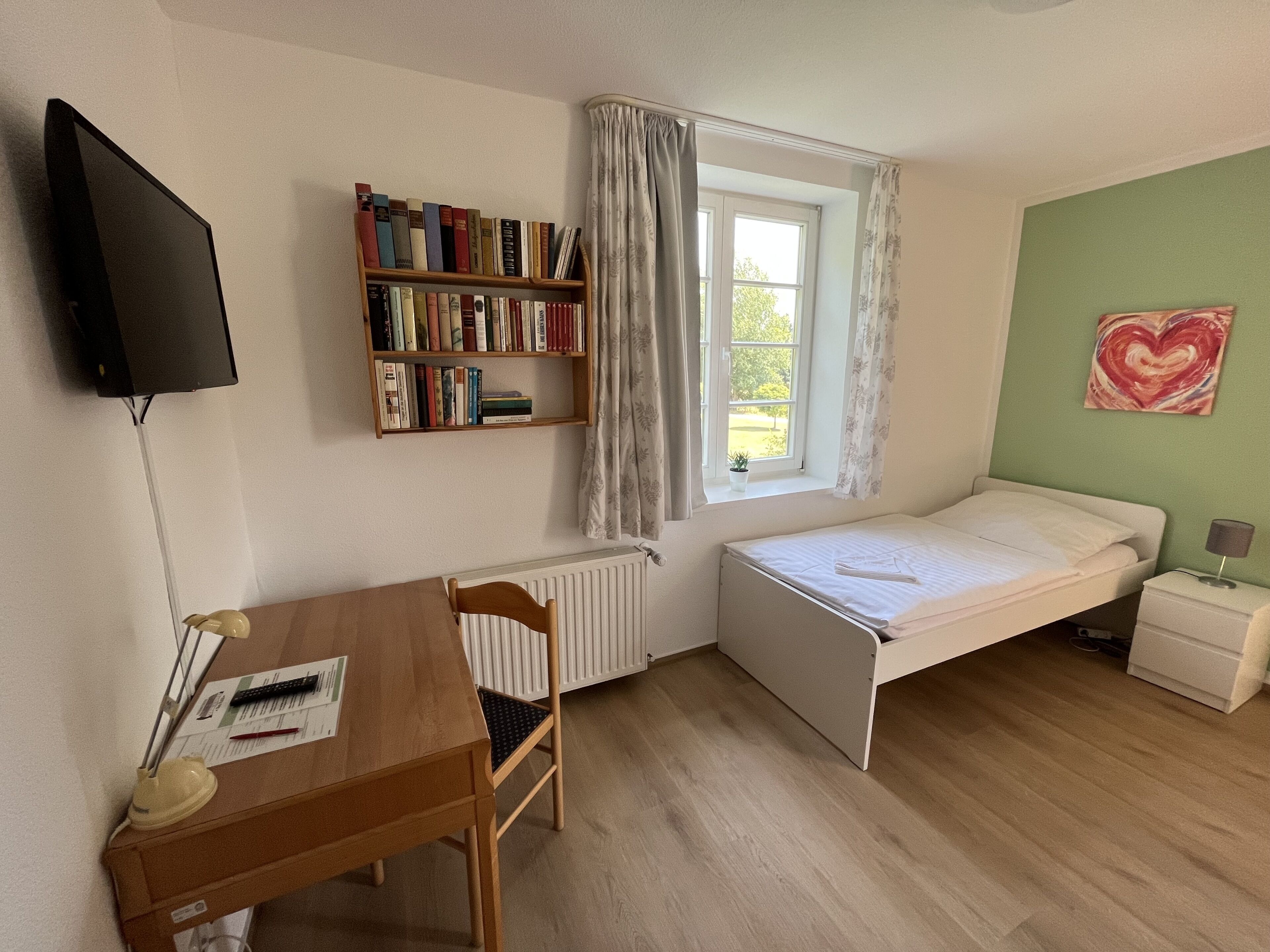 Una habitación pequeña con cama, escritorio, estantería, radiador, ventana con cortinas y un corazón decorativo en la pared.