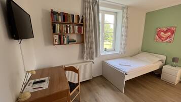 Una habitación pequeña con cama, escritorio, estantería, radiador, ventana con cortinas y un corazón decorativo en la pared.