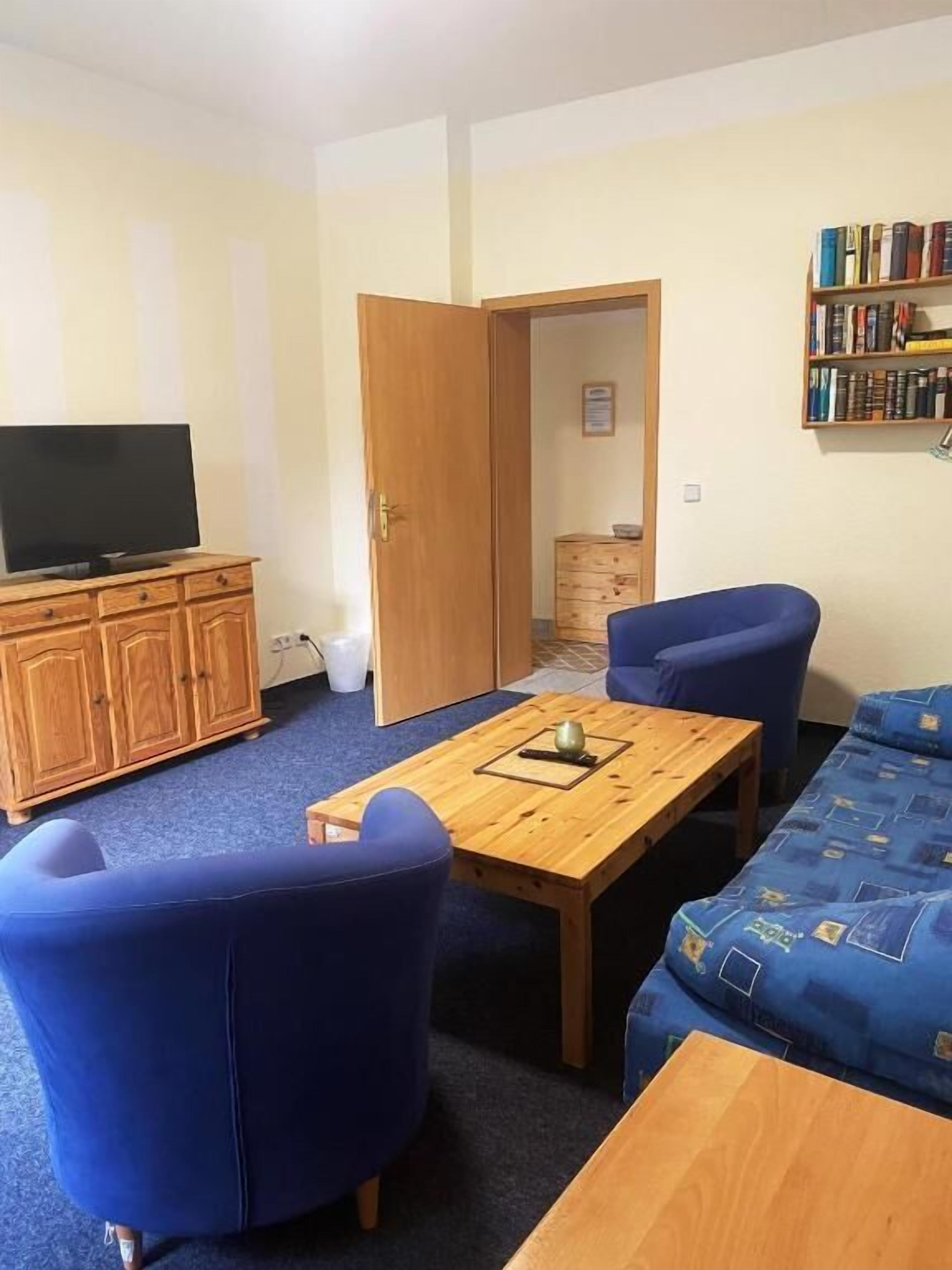 A room with a television on a wooden cabinet, a table with a book and a cup, blue chairs, a sofa, and a bookshelf.