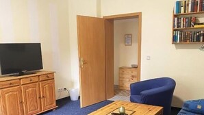 Una habitación con una televisión sobre un mueble de madera, una mesa con un libro y una taza, sillas azules, un sofá y una estantería.