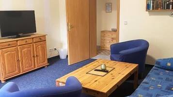 A room with a television on a wooden cabinet, a table with a book and a cup, blue chairs, a sofa, and a bookshelf.