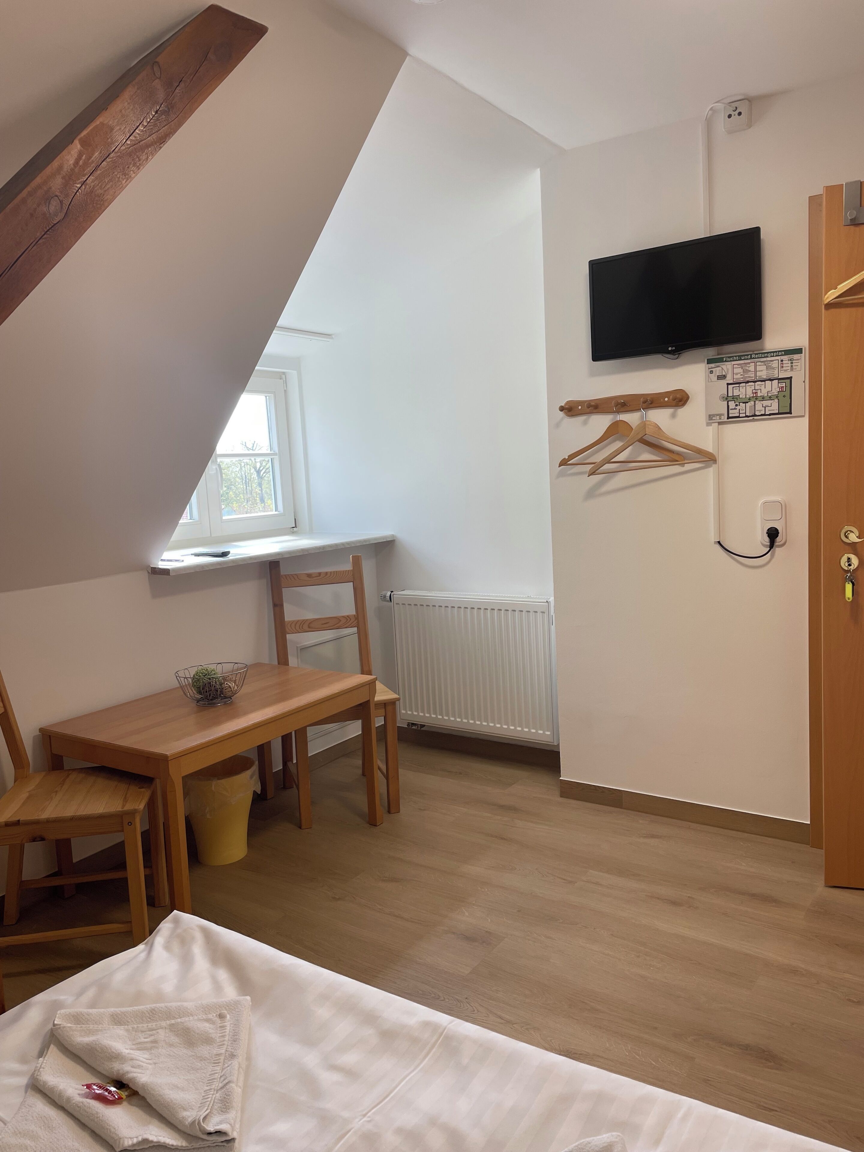 A room with a sloped ceiling, a wooden table with chairs, a bed, a radiator, a TV mounted on the wall, and a door with a handle.