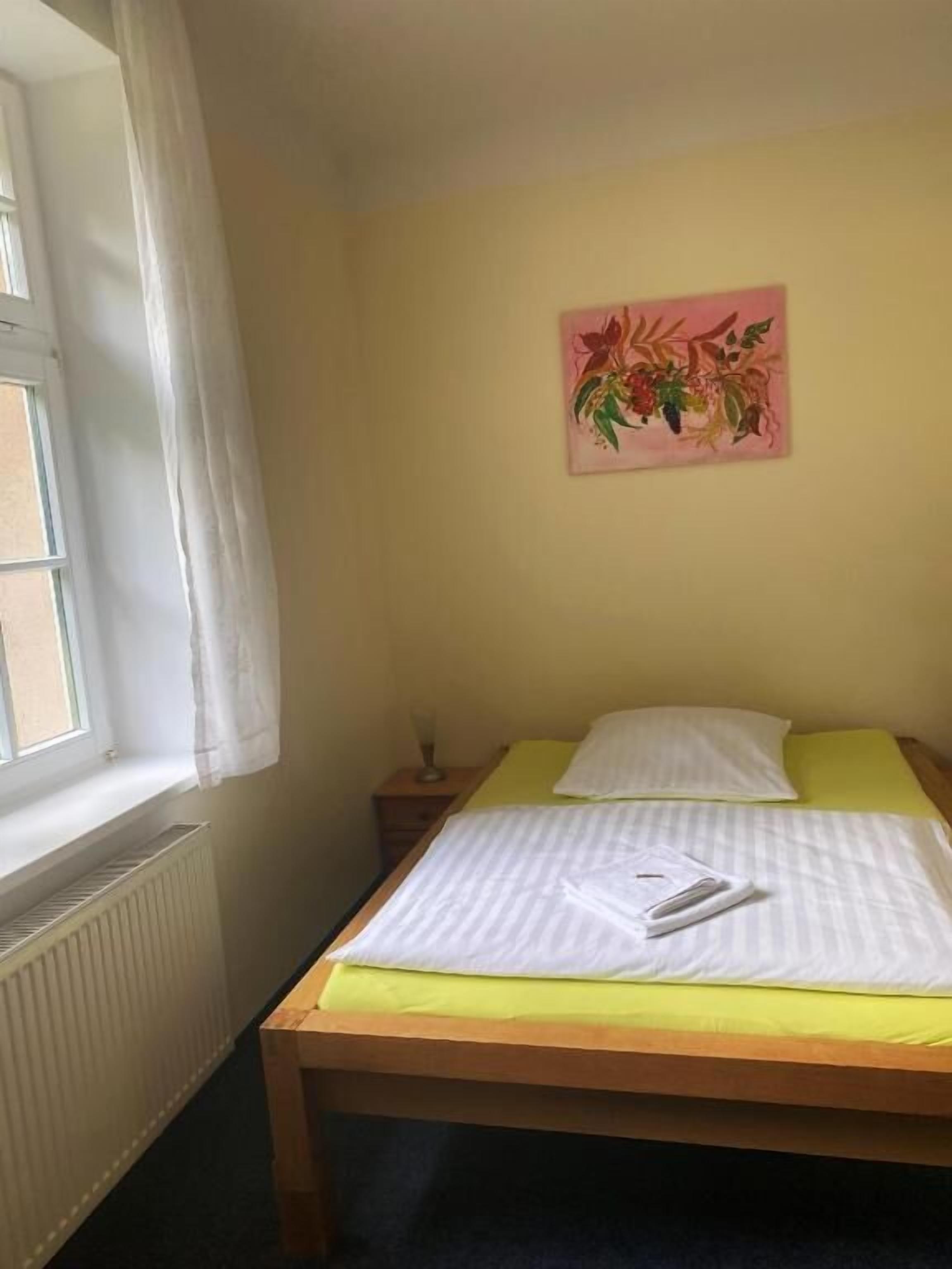 A single bed with a yellow bedspread, a white pillow, and a white blanket. There is a nightstand with a glass and a lamp. A window with a white curtain is visible, and a painting of flowers hangs on the wall.