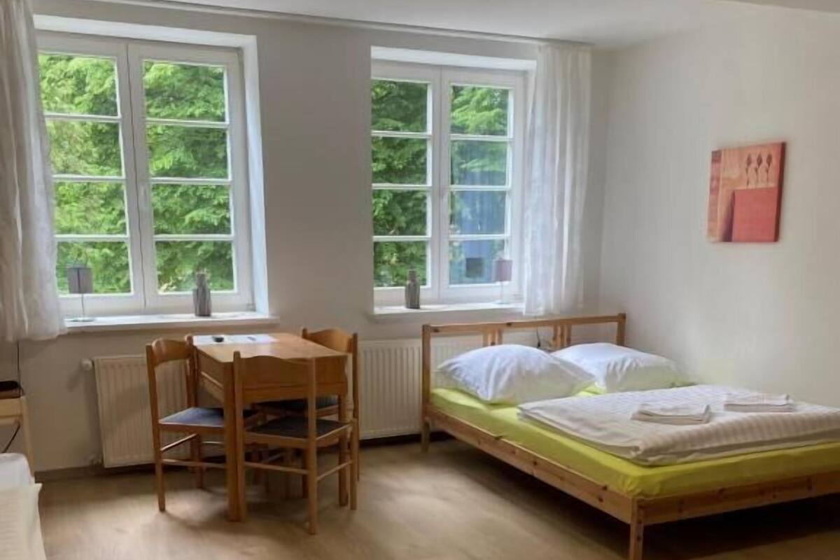 A room with a bed, a wooden table, and two windows with curtains.