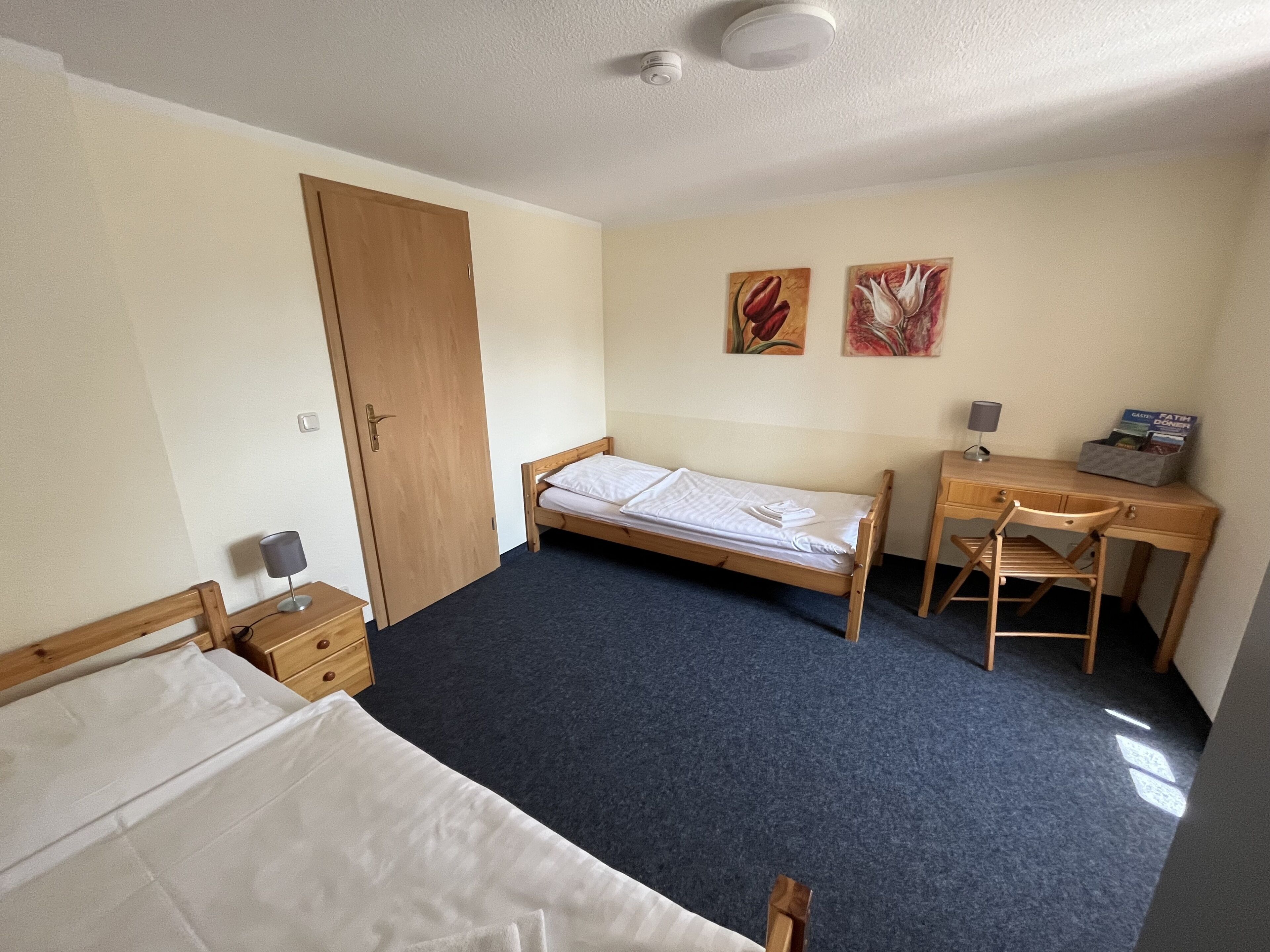 Une chambre individuelle comprenant un lit, un bureau, une chaise, une armoire et deux tableaux accrochés au mur.
