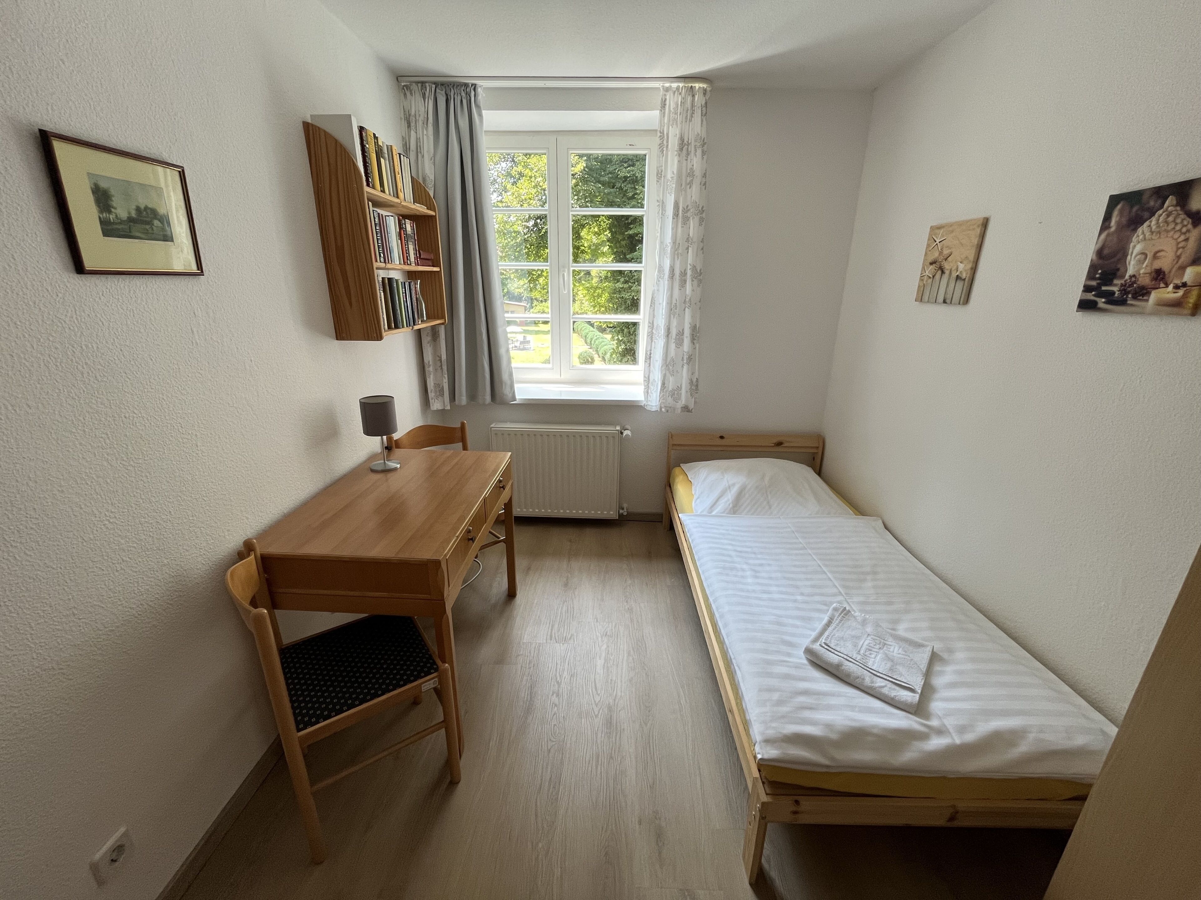 A small, well-lit room with a wooden desk, a single bed, a radiator, and a window with curtains.