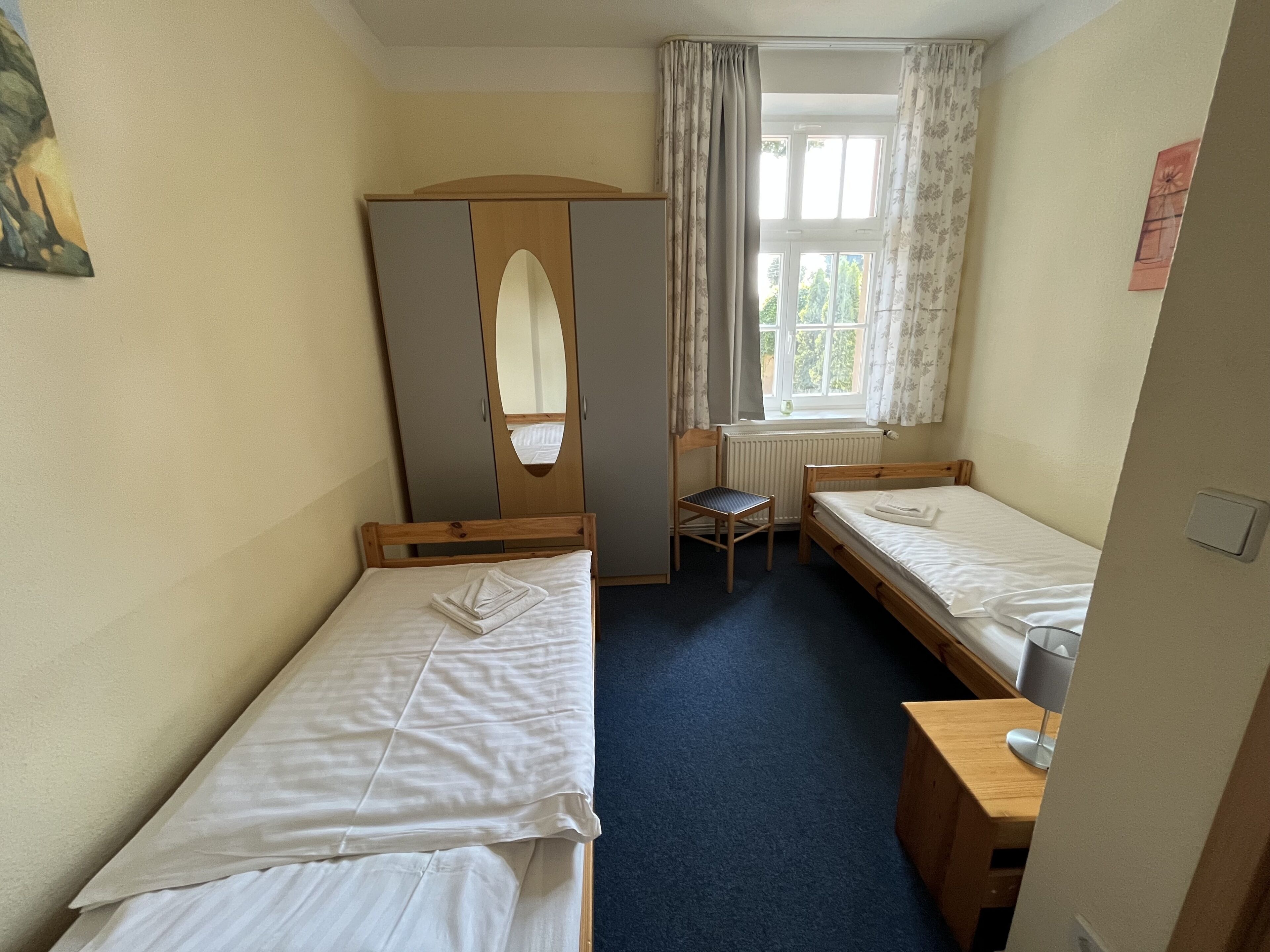 Habitación de hotel con dos camas individuales, un armario de madera, una silla, una mesita y una ventana con cortinas.