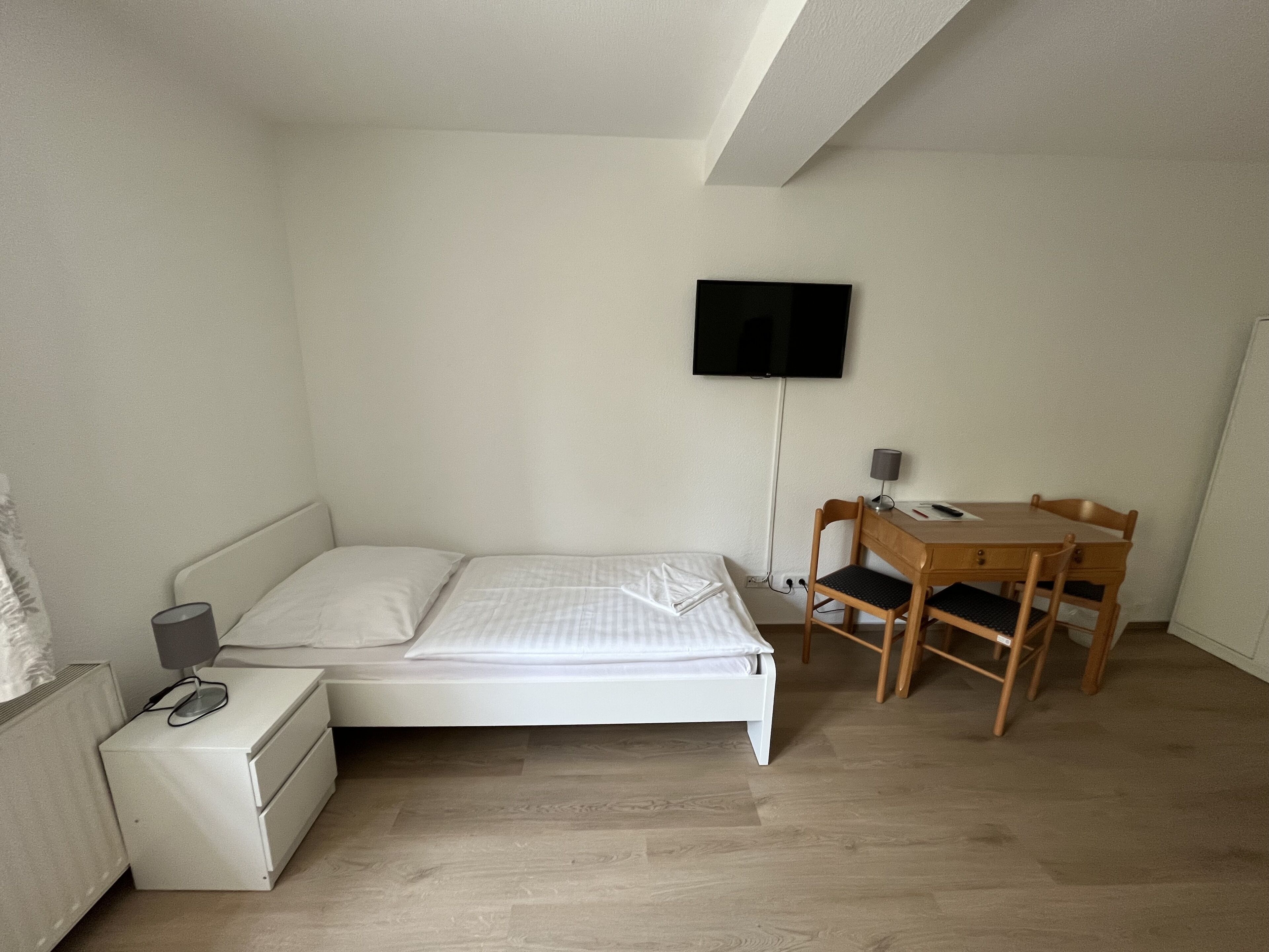 Ein kleines, ordentlich eingerichtetes Schlafzimmer mit einem Einzelbett, einem Nachttisch mit Lampe, einem Holztisch mit Stuhl und einem an der Wand montierten Flachbildfernseher.