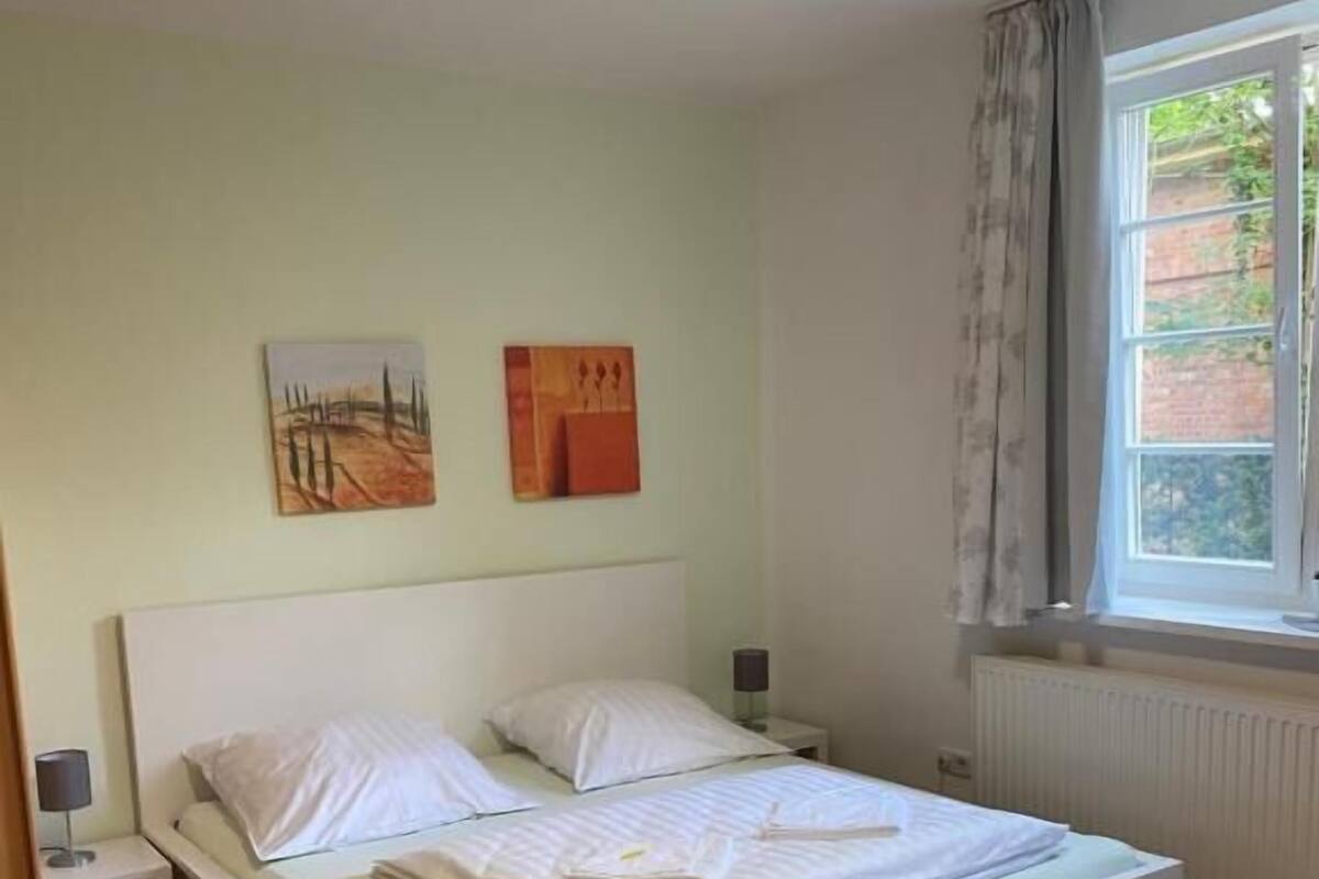 A single-bed room with a white headboard, a nightstand, a lamp, and two wall-mounted paintings.