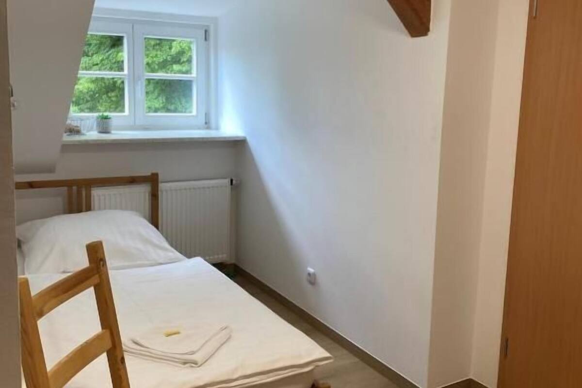 A bedroom with a sloped ceiling, a window, a radiator, a wooden bed frame, a wooden chair, and a wooden door.