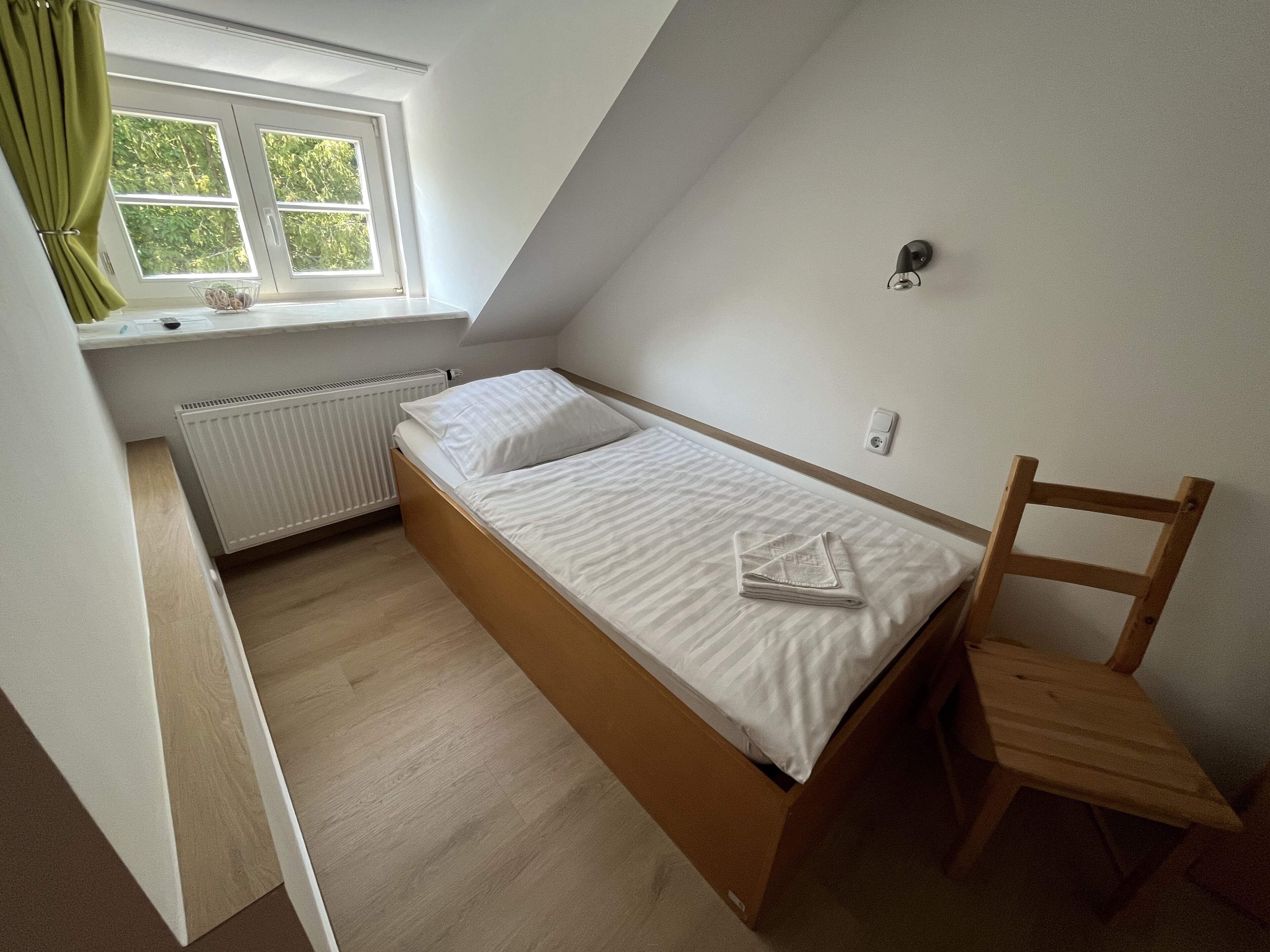 A small, single-bed room with a wooden frame, a window with curtains, a radiator, and a chair.