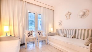 Una habitación con sofá, dos sillones con cojines, lámpara y vistas al exterior.