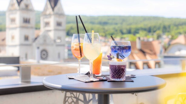 Una mesa con tres bebidas en un balcón con vistas a una ciudad y una torre de iglesia.