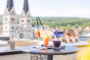Une table avec trois boissons sur un balcon donnant sur une ville, avec le clocher d’une église en arrière-plan.