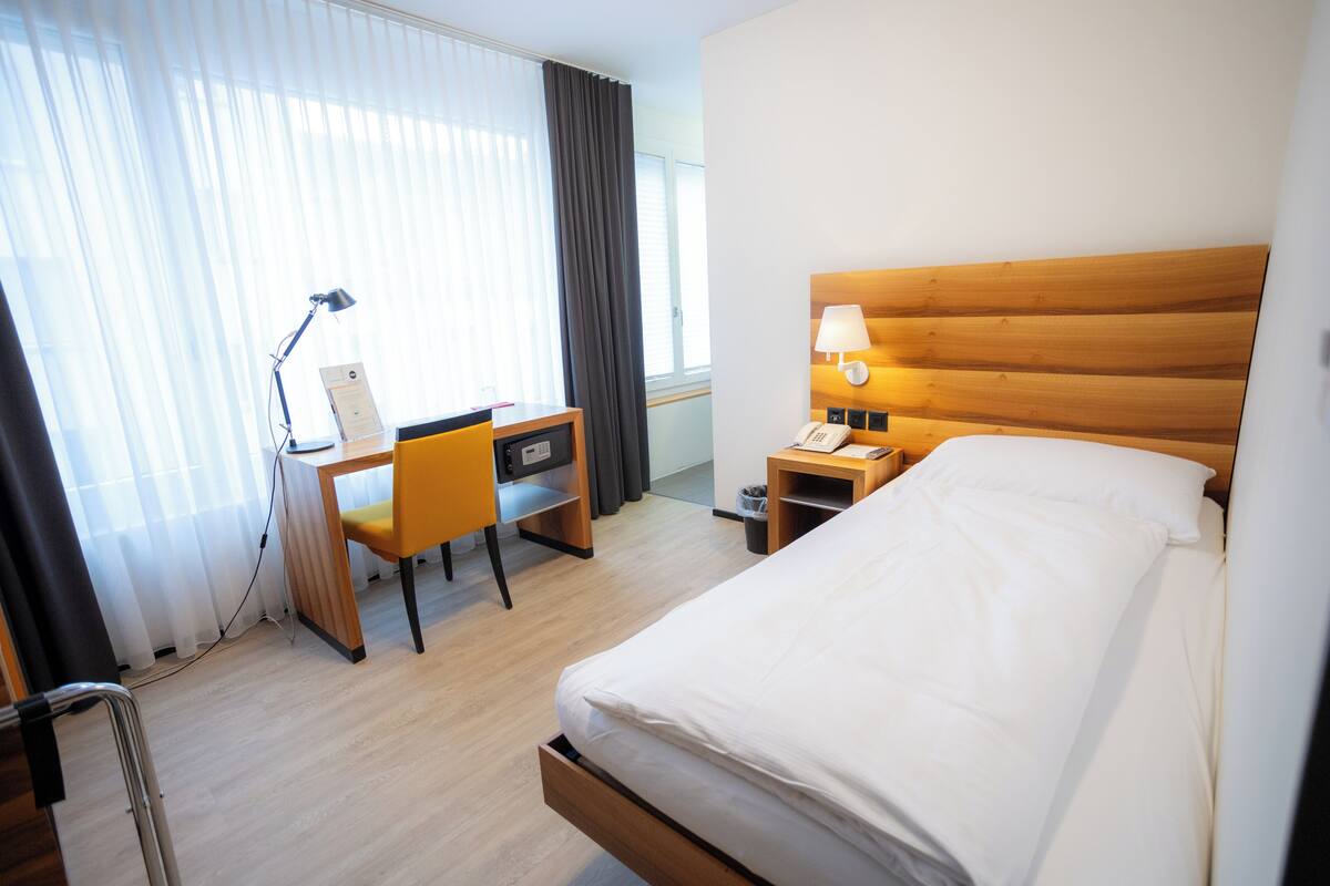 Una habitación de hotel con cama, escritorio, silla, lámpara y ventana con cortinas.