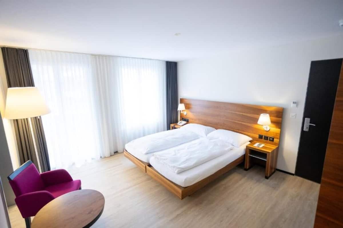 Habitación de hotel con cama, dos lámparas de noche, una mesita de noche, una silla y una mesa.