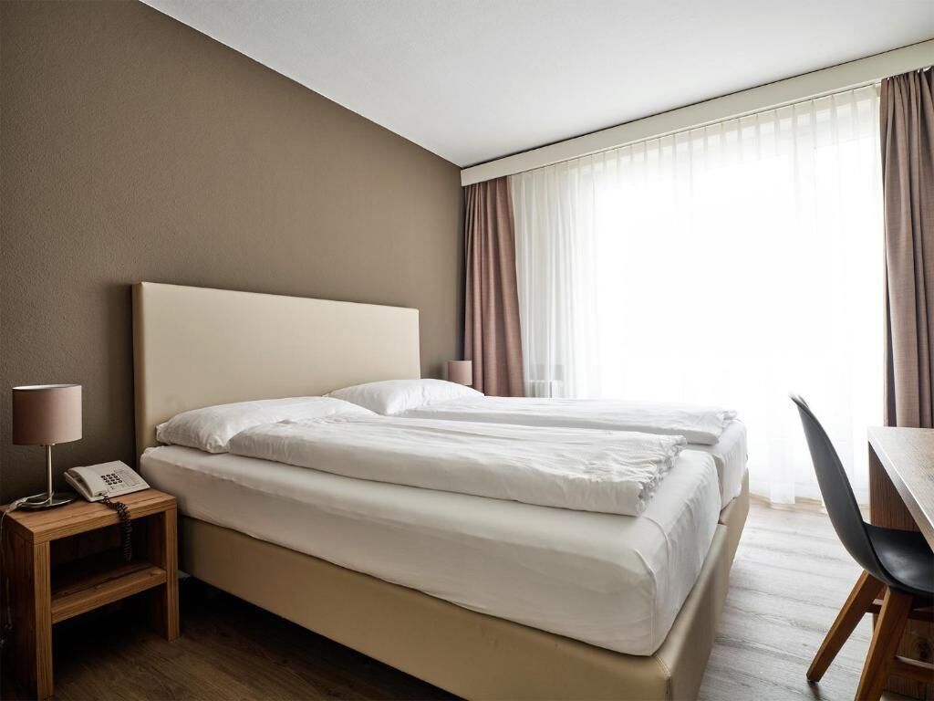 Habitación de hotel con cama, mesita de noche con lámpara y teléfono, escritorio con silla y ventana con cortinas.