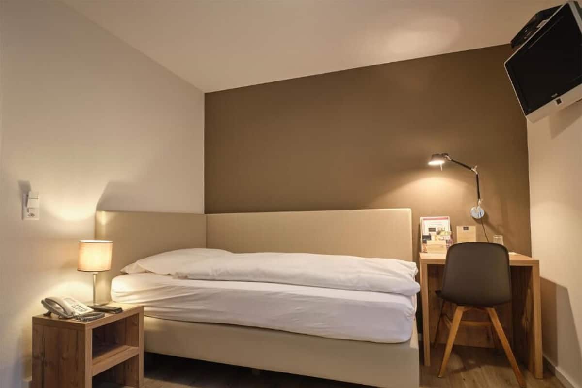 Habitación de hotel con cama, mesita de noche con lámpara, escritorio con silla y televisor de pantalla plana montado en la pared.