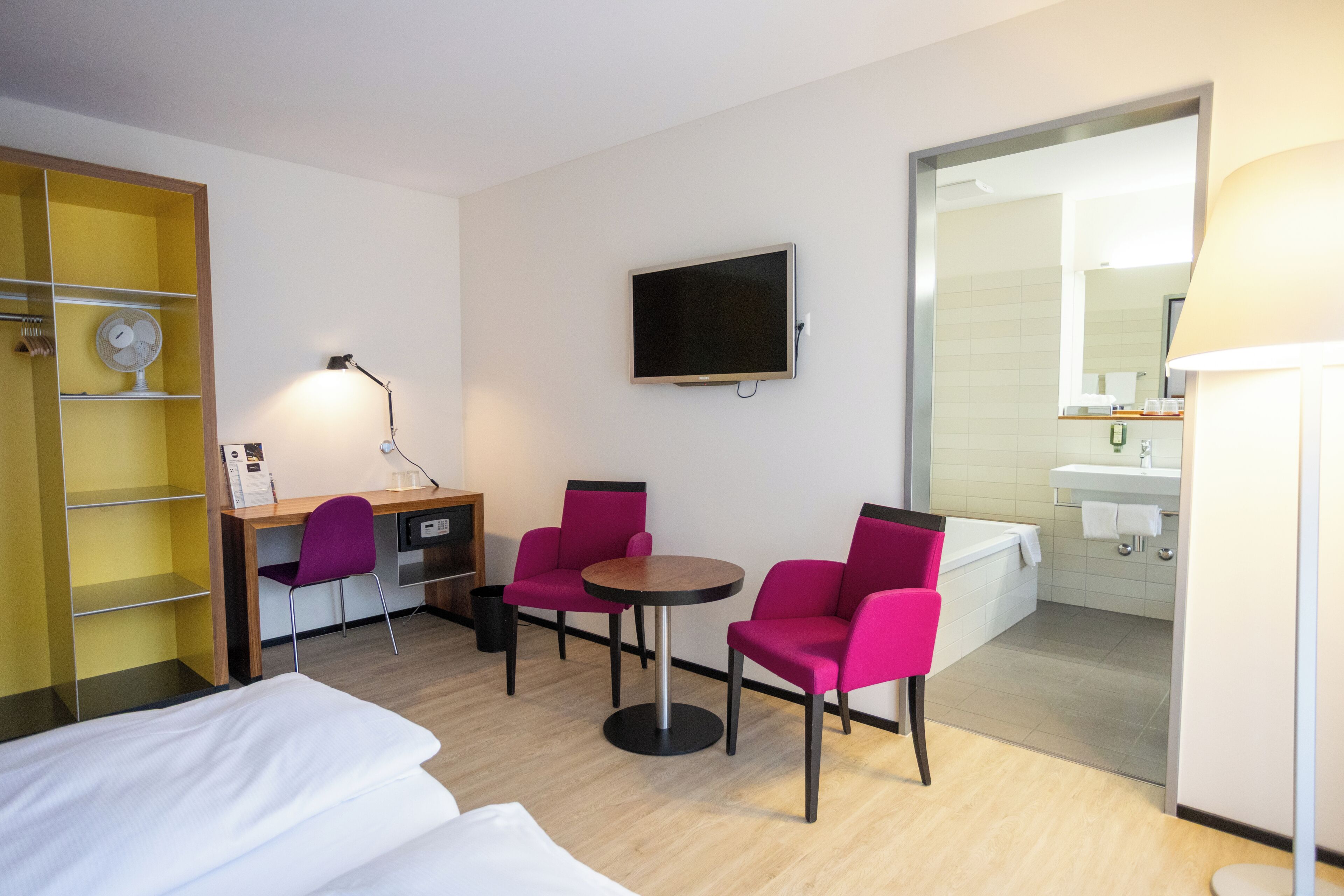 Ein Hotelzimmer mit einem Bett, einem Schreibtisch, zwei Stühlen, einem Fernseher, einer Lampe, einem Spiegel und einem durch eine offene Tür einsehbarer Badezimmer.