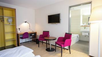 Ein Hotelzimmer mit einem Bett, einem Schreibtisch, zwei Stühlen, einem Fernseher, einer Lampe, einem Spiegel und einem durch eine offene Tür einsehbarer Badezimmer.