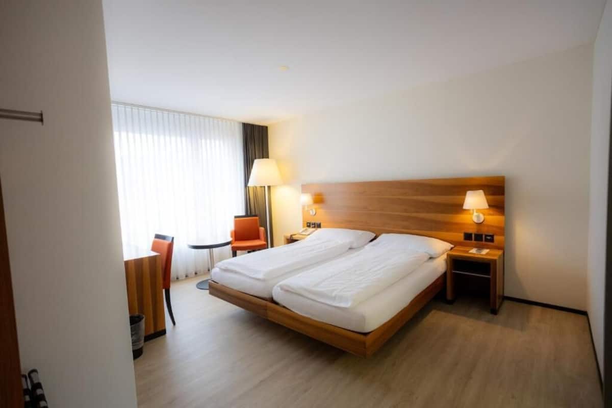Habitación de hotel con cama doble, dos mesitas de noche con lámparas, una mesita pequeña con una silla y una ventana con persianas.