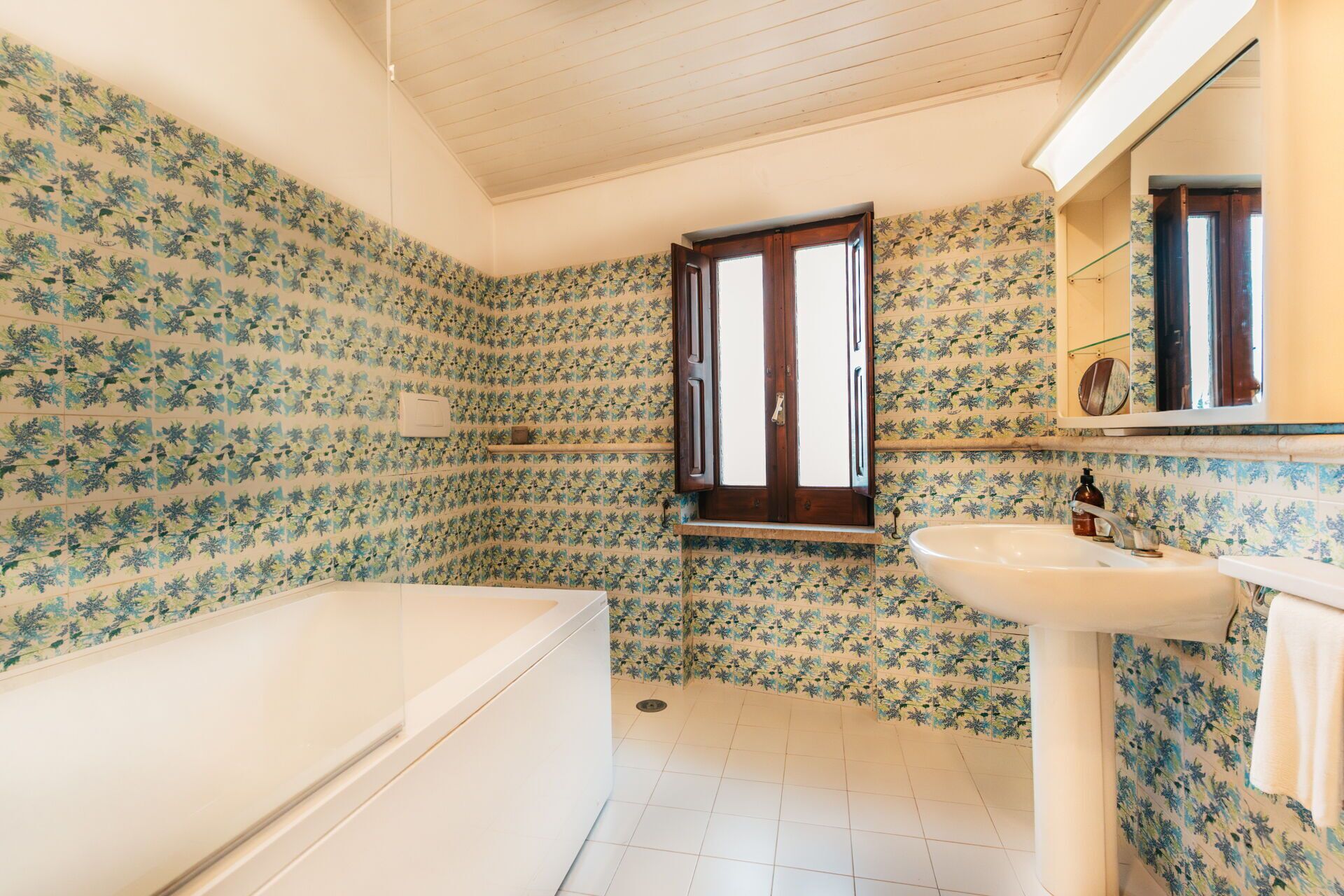 A bathroom with floral patterned tiles, a bathtub, a sink, and a window with wooden shutters.