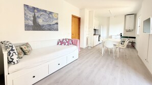 A modern room with a white sofa, a painting on the wall, a dining area with a table and chairs, and a kitchenette in the background. - M462 - Marcelli, three-room apartment near the sea (Marcelli)