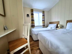 Habitación de hotel con dos camas, una silla, una mesita de noche con lámpara y una ventana con cortinas.