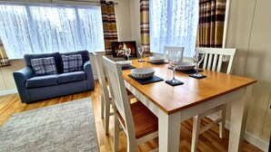 A dining area with a wooden table, white chairs, and a blue sofa. - Coastal Bella Mere Lodge • 2 Bed • Sleeps 5 (Ilfracombe)
