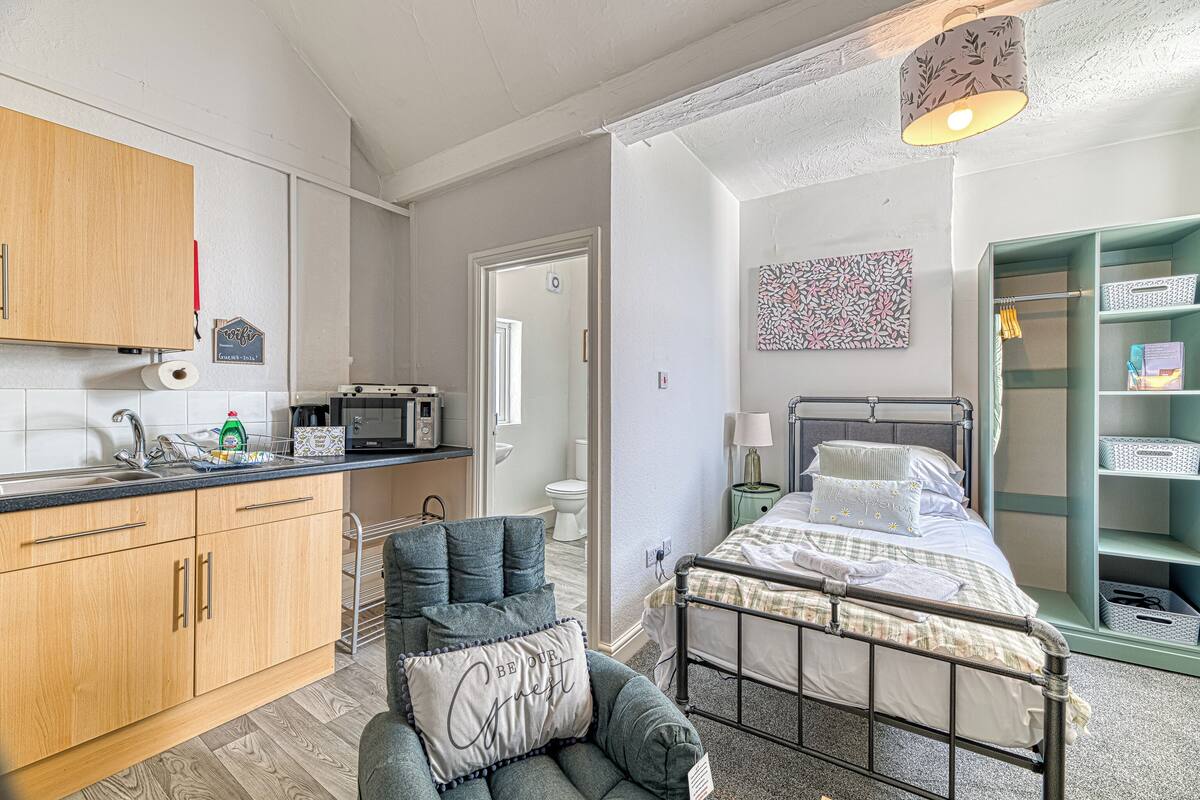 A compact room with a kitchenette, a bed with a headboard, a bedside table, and a green shelving unit.