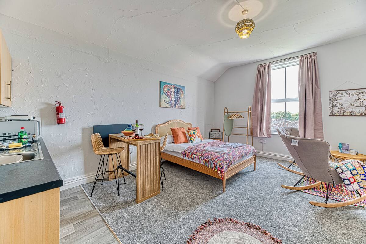 A bedroom with a bed, a desk, a rocking chair, a window with curtains, and a fire extinguisher.