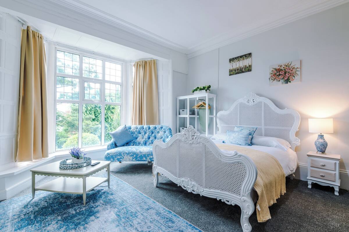 A bedroom with a white bed, a blue sofa, a white nightstand, a white table, and a window with curtains.