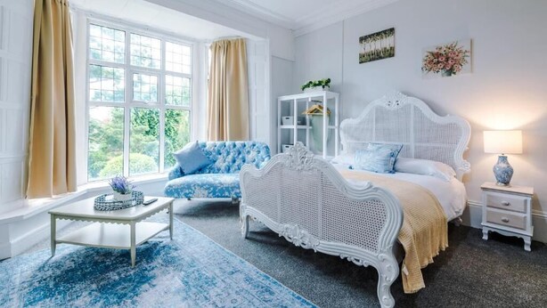 Una habitación con una cama blanca, un sofá azul, una mesita de noche blanca y una ventana con cortinas.