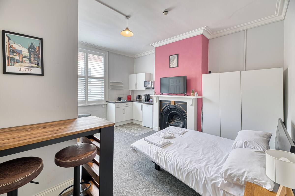 A bedroom with a bed, a desk with stools, a television, a fireplace, and a kitchen area.