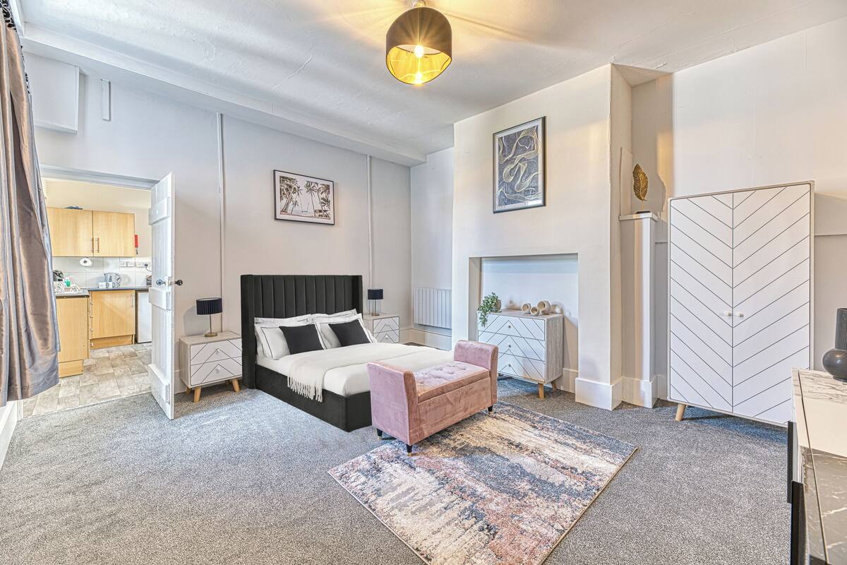 A bedroom with a bed, bedside tables, a dresser, a fireplace, and a kitchen in the background.