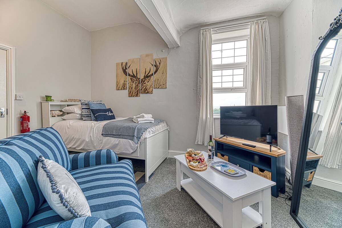 A room with a bed, a TV, a sofa, a small table with food, and artwork on the wall.