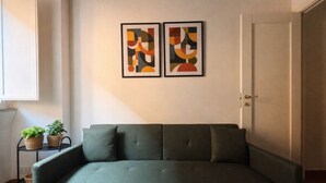 Huoneessa on vihreä sohva, ovi, pieni pöytä, jossa on koreja, sekä kaksi abstraktia maalausta seinällä.