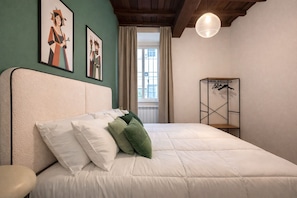 Ein Schlafzimmer mit Bett, Kopfteil, einer Hängeleuchte, einem Fenster mit Vorhängen und einem an der Wand befestigten Kleiderhaken.
