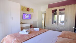 Ein Schlafzimmer mit Bett, Fernseher, Kleiderschrank und einem durchgehenden Türrahmen sichtbaren Badezimmer.