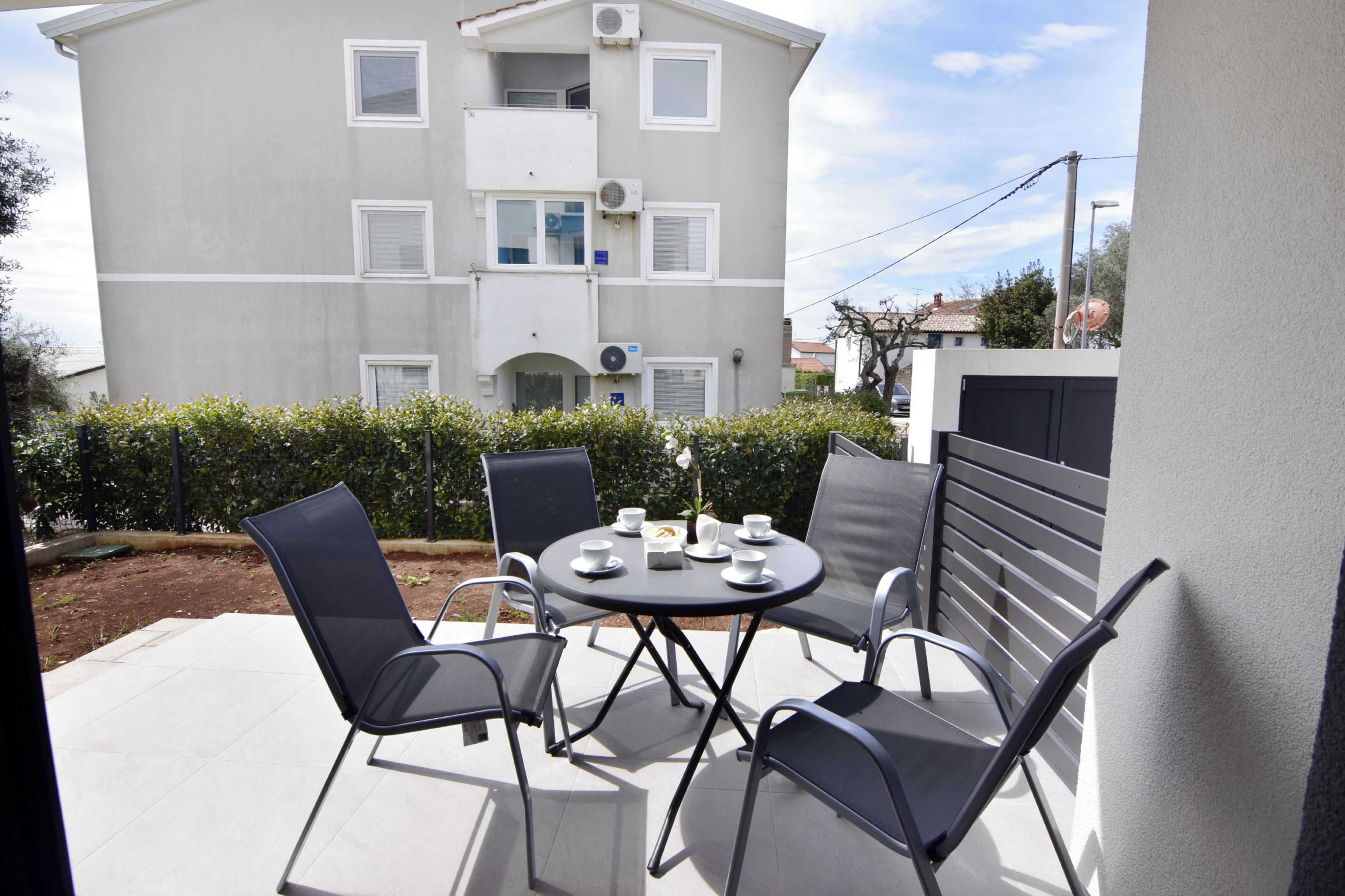 Een terras buiten met een ronde tafel en vier stoelen, een klein tafeltje met kopjes en een bloem, en een gebouw met meerdere ramen en balkons op de achtergrond.