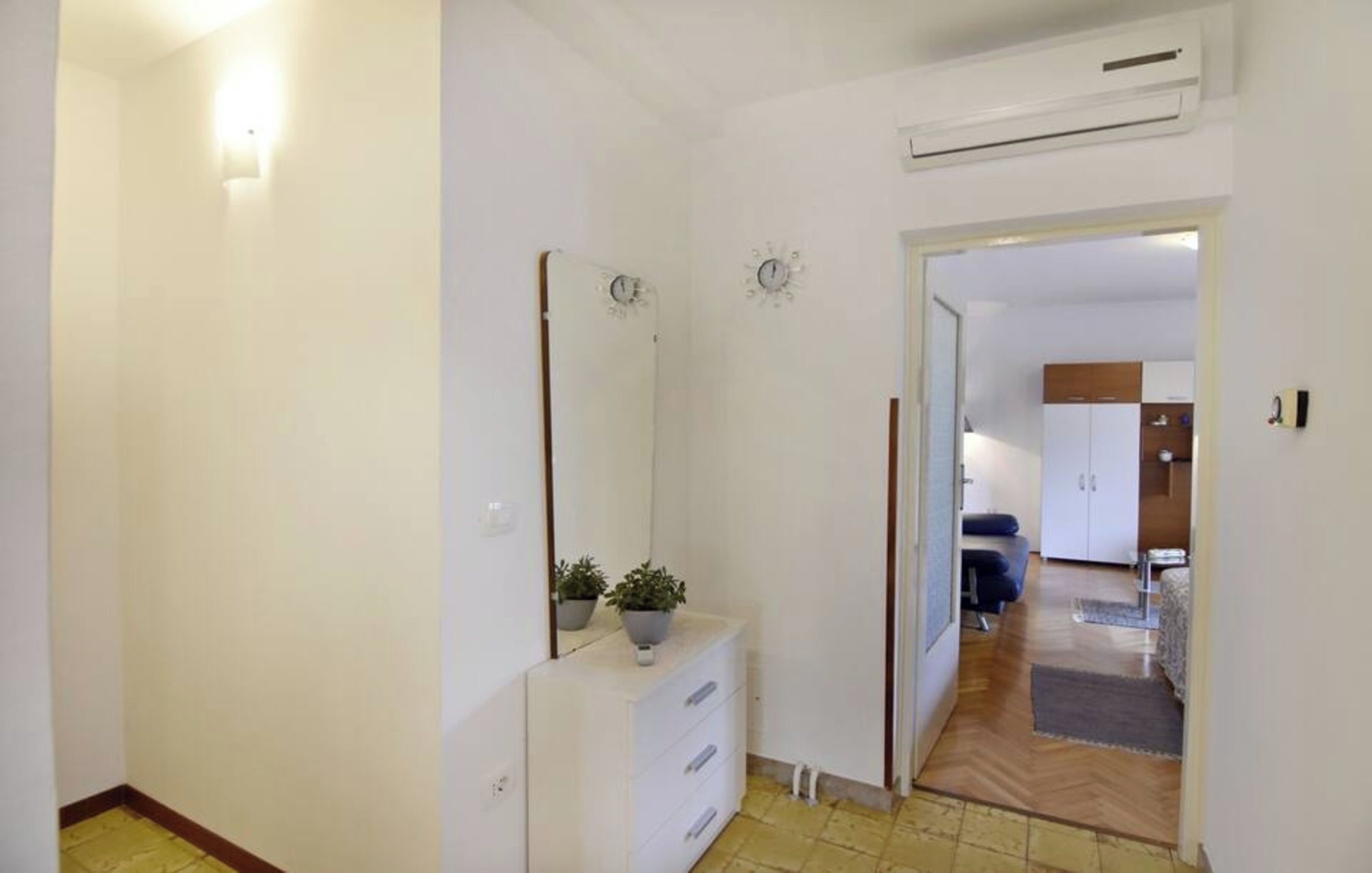 Un pasillo con un aparador blanco, un espejo y una planta en maceta. Se ve una puerta que conduce a una habitación con cama y armario.