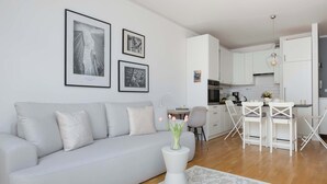 A modern living room with a sofa, a coffee table with a vase, and a kitchen area with white cabinets and a dining set. - Modern Apartment | Balcony and Workspace (Warszawa)
