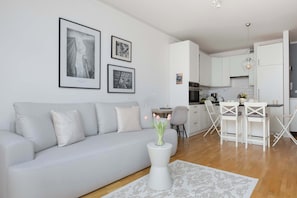 A modern living room with a sofa, a coffee table with a vase, and a kitchen area with white cabinets and a dining set. - Modern Apartment | Balcony and Workspace (Warszawa)
