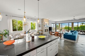 A modern kitchen with a large island, dining area with white furniture, and a living room with a blue sofa. - Casa Solara: Panoramic Views, Pool & Spa, GameRoom (Desert Hot Springs)