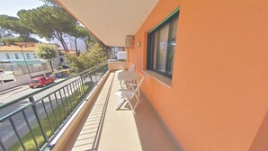 A balcony with a table and chairs, a view of the street, and a building with an orange facade. - Bright holiday flat close to the beach (Lignano Sabbiadoro)