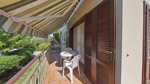 En balkon med et bord og stole, en trædør og udsigt til grøn vegetation.
