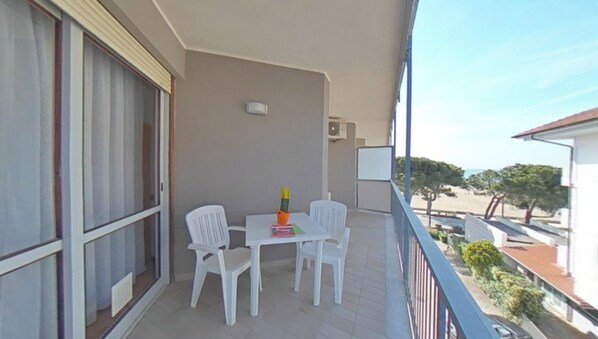 A balcony with a table and chairs, a window with blinds, and a view of the beach and buildings. - Prime beachside apartment (Lignano Sabbiadoro)