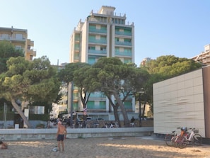 Une plage de sable avec un grand immeuble d’habitation en arrière-plan, des vélos garés près d’un bâtiment et un ciel dégagé.