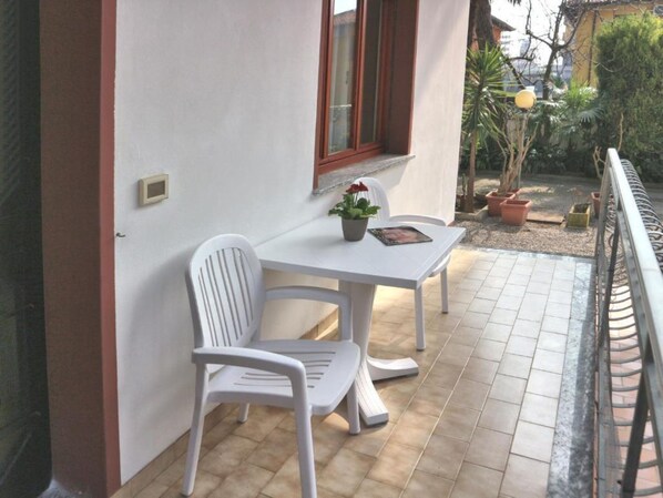 Een klein buitenterras met een witte tafel en stoelen, een bloempot en een raam met een rode omlijsting.