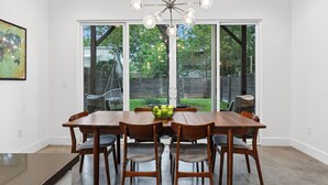 Un comedor con mesa y sillas de madera, una araña y un amplio ventanal corredera que da a un jardín.